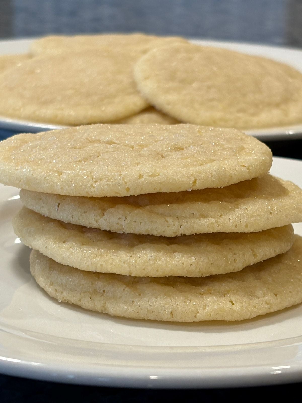 A stack of five lemon sugar cookies on a white plate, with more cookies in the background. The cookies look soft and lightly sprinkled with sugar, making this an irresistible lemon cookie recipe for any sweet tooth.