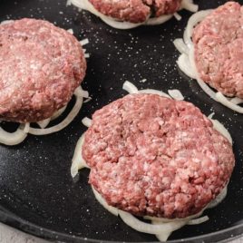 close up shot of uncooked Oklahoma Fried Onion Burgers over sliced onions in a skillet