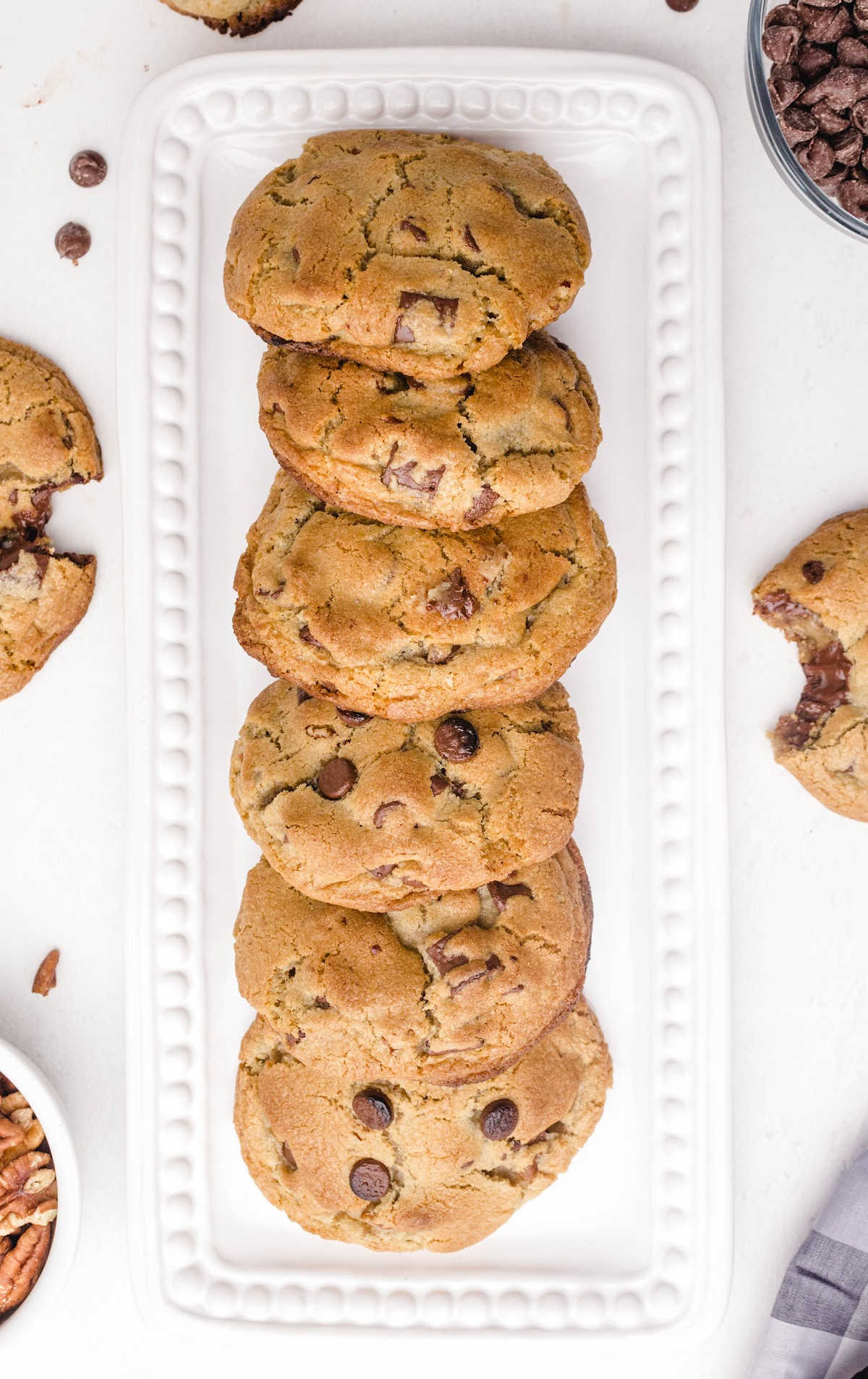 Overhead view of thick chocolate chip cookies neatly stacked on a white rectangular plate with chocolate chips and pecans scattered around.
