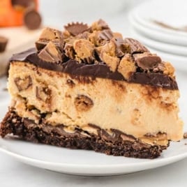 Close-up of a single slice of Reese’s cheesecake on a white plate, loaded with chunks of peanut butter cups.