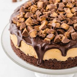 close up shot of Reese’s cheesecake topped with chocolate ganache and pieces of Reese's cups on a cake serving tray