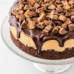 close up shot of Reese’s cheesecake topped with chocolate ganache and pieces of Reese's cups on a cake serving tray