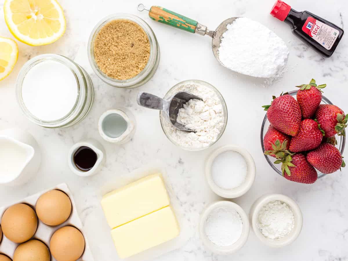 Baking ingredients on a marble surface: eggs, butter, strawberries, sugar, brown sugar, flour, lemon slices, vanilla, milk, baking powder, powdered sugar, salt, and a bottle of extract—everything you need to create the perfect Strawberry Lemon Blondies Recipe.