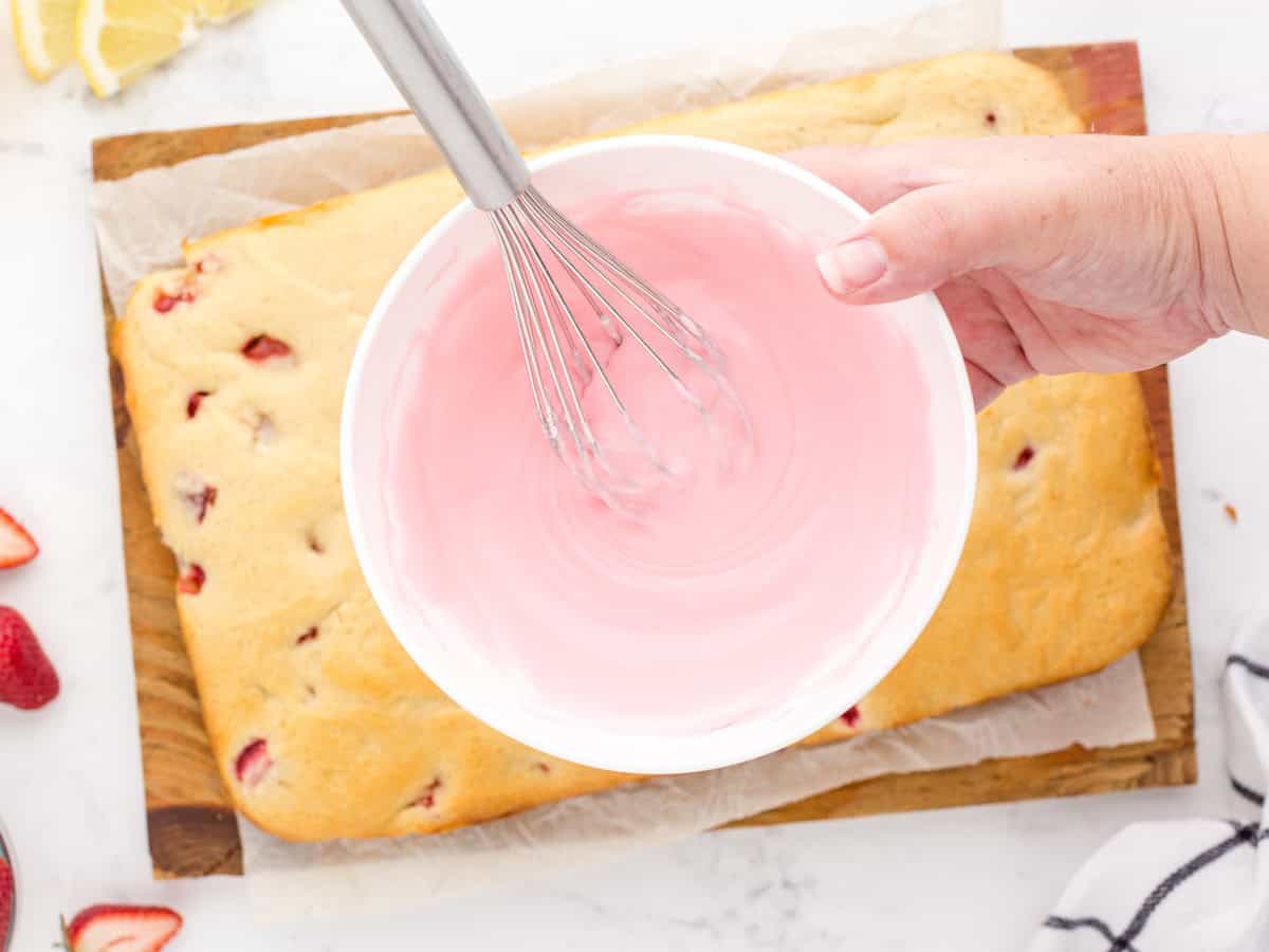 A hand holds a whisk and mixes pink icing in a white bowl above a rectangular strawberry cake on parchment paper, preparing the finishing touch for these irresistible Strawberry Lemon Blondies.
