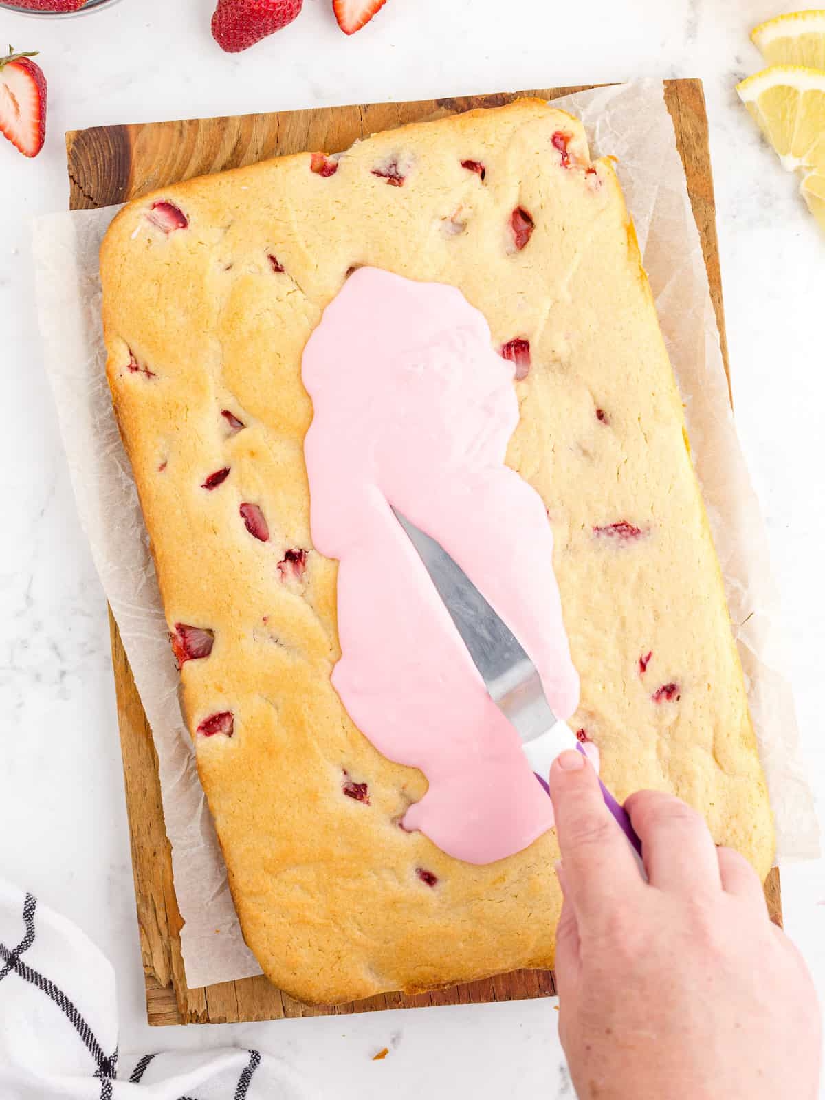 A hand spreads pink frosting on a rectangular strawberry cake with a spatula, preparing delicious Strawberry Lemon Blondies. The cake rests on parchment paper atop a wooden board, with sliced strawberries and lemon nearby, making this Strawberry Lemon Blondies Recipe irresistibly fresh and vibrant.