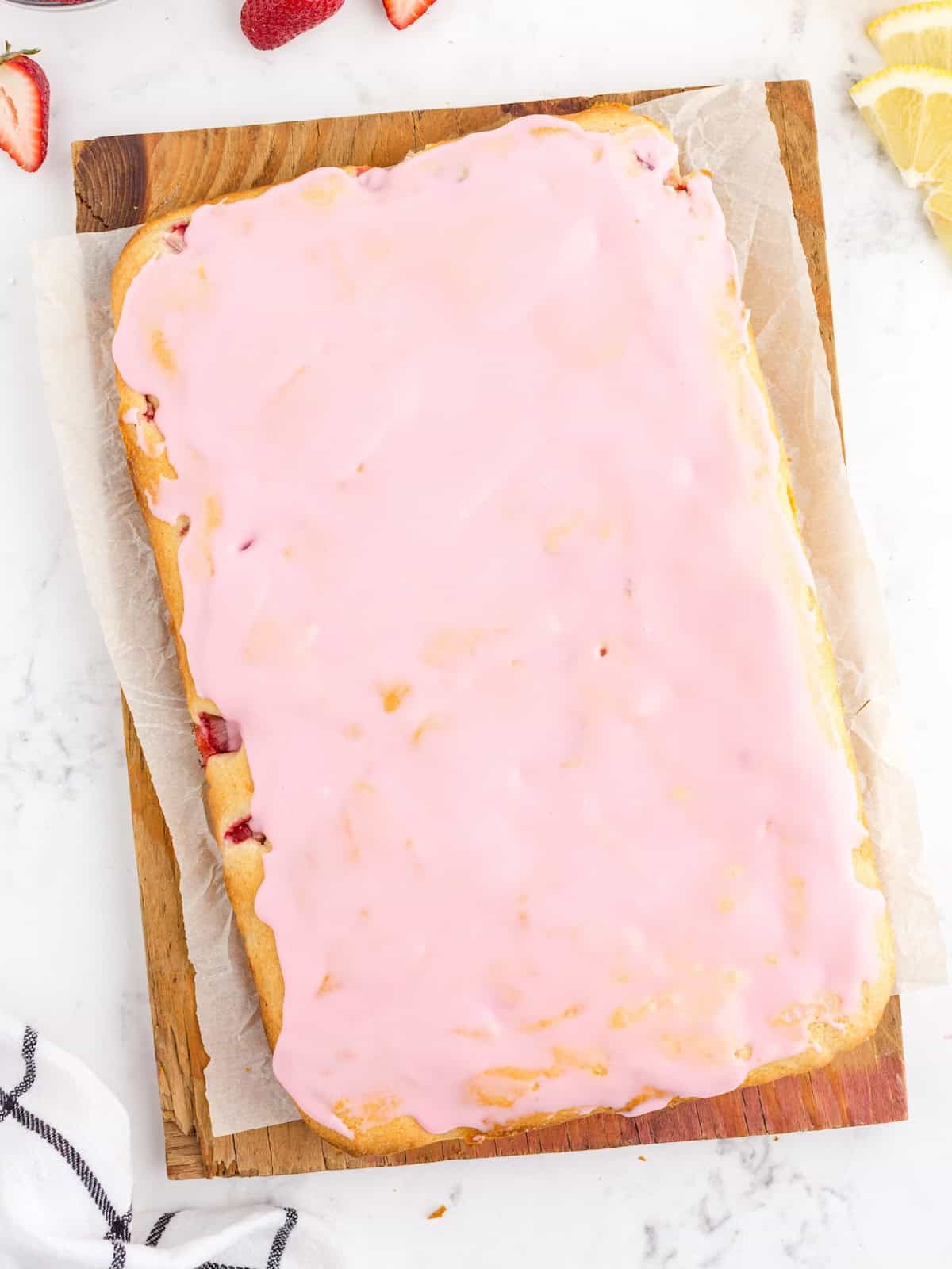 A rectangular sheet cake with pink icing sits on parchment paper atop a wooden board. Slices of lemon and strawberries are visible nearby on a white marble surface, making this dessert the perfect centerpiece for any Strawberry Lemon Blondies Recipe.