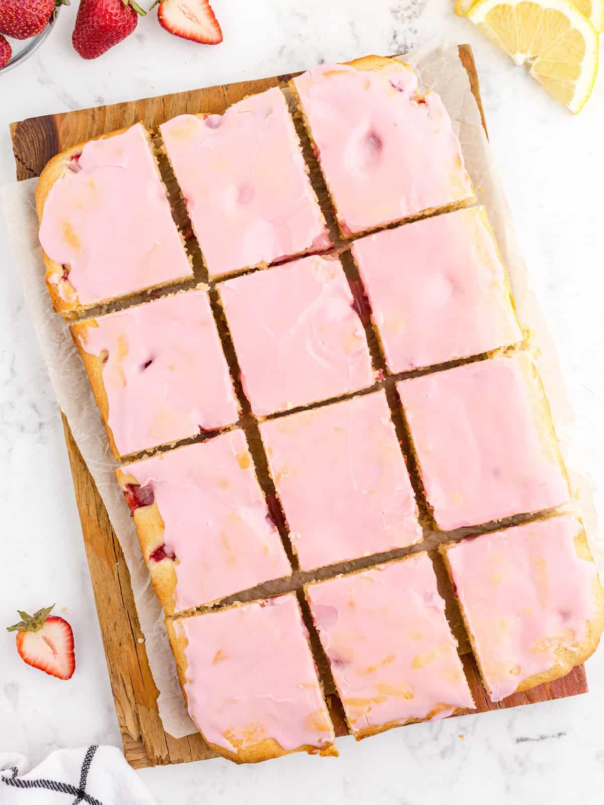 Twelve rectangular Strawberry Lemon Blondies with pink icing are arranged on a parchment-lined wooden board. Fresh strawberries and a lemon slice are nearby on a marble surface, showcasing the vibrant ingredients in this delicious Strawberry Lemon Blondies recipe.