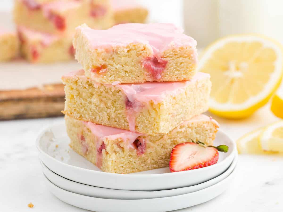Three lemon-strawberry bars with pink icing, inspired by a Strawberry Lemon Blondies recipe, are stacked on white plates. A sliced strawberry and lemon are beside them, while more Strawberry Lemon Blondies are blurred in the background.