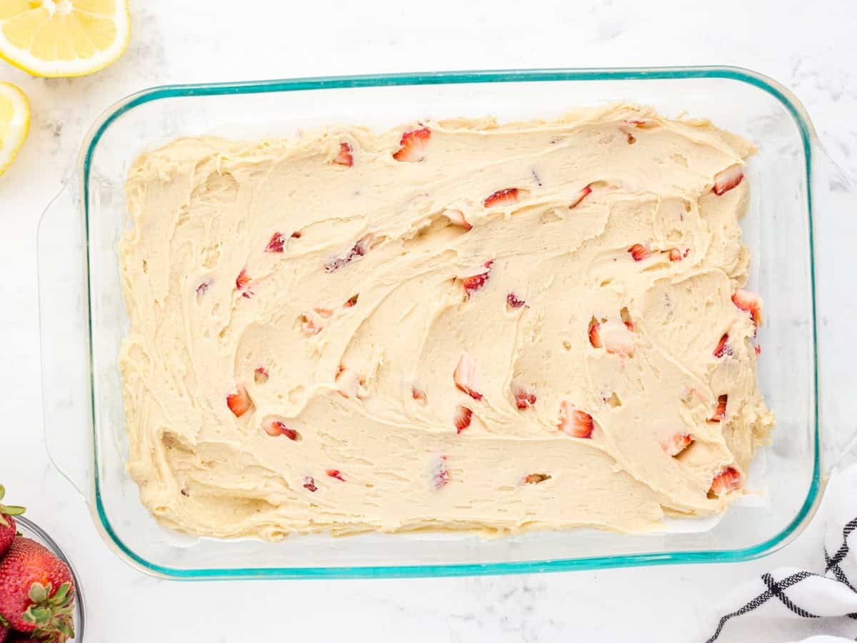 Rectangular glass baking dish filled with unbaked Strawberry Lemon Blondies batter, mixed with strawberry pieces, on a white surface with lemon slices and strawberries partially visible nearby—perfect for making your favorite Strawberry Lemon Blondies Recipe.