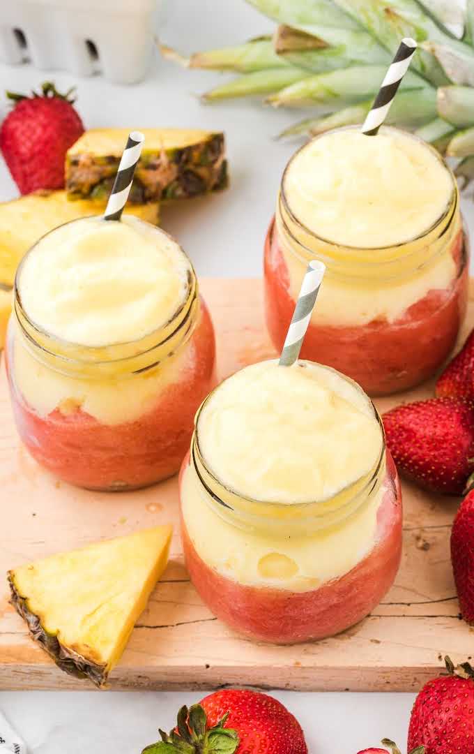 close up shot of wine slushies in mason jars with strawberries and pineapples with straws on a wooden board