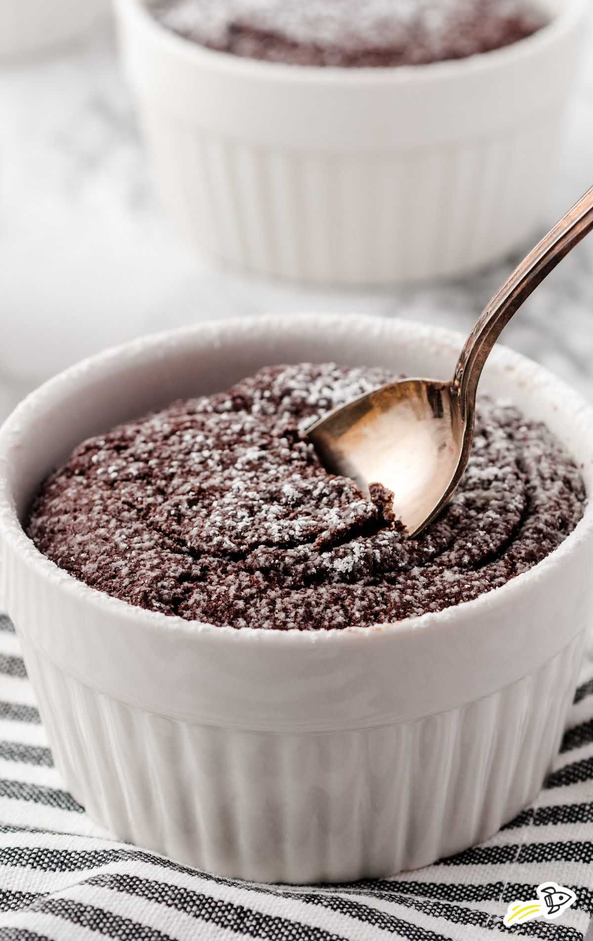 Baked chocolate lava cake topped with powdered sugar being spooned from a white ramekin on striped cloth.