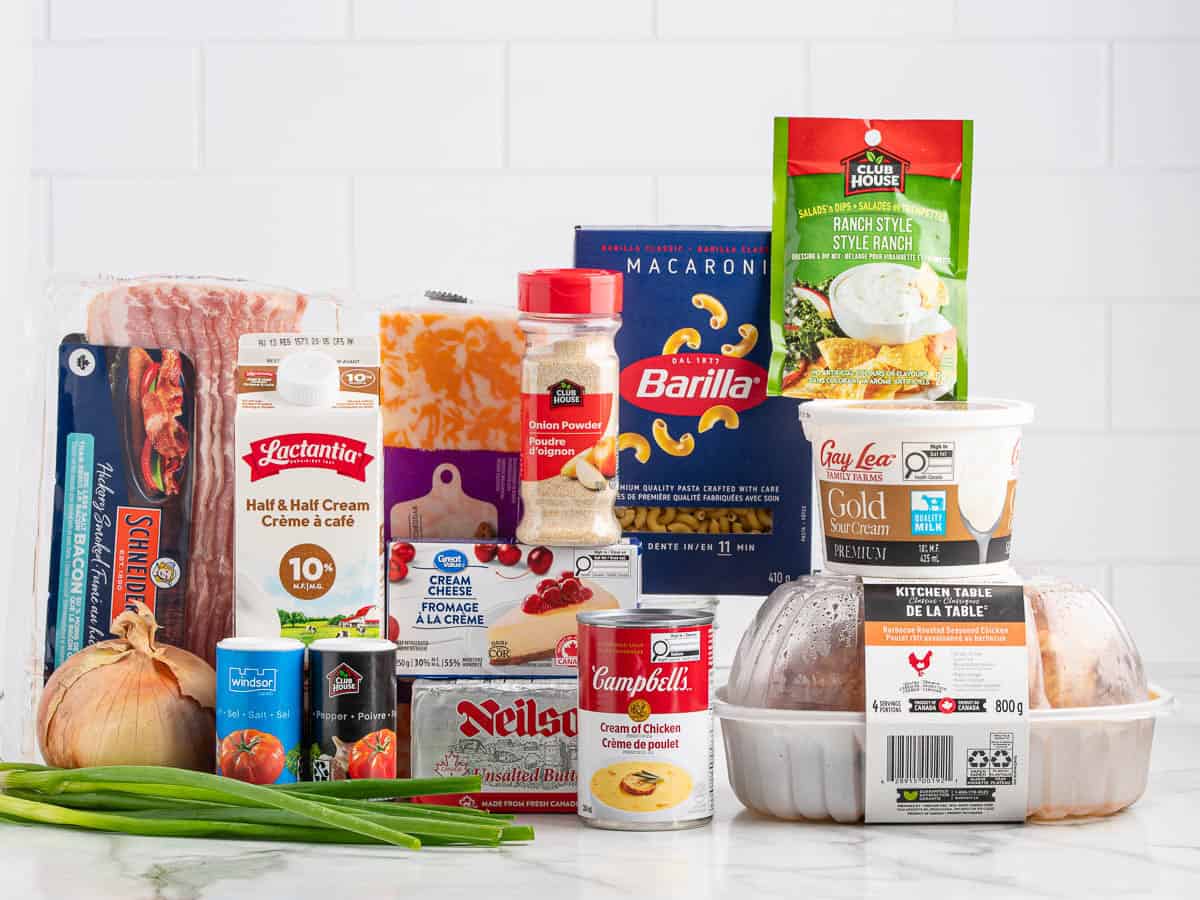 Assorted groceries, perfect for making a delicious Crack Chicken Casserole Recipe, including pasta, cream, cheese, bacon, soup, green onions, an onion, a rotisserie chicken, and other pantry items on a kitchen counter with a white tile background.