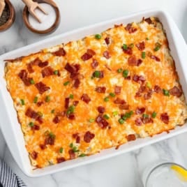 A casserole dish filled with baked cheesy hash browns, topped with melted cheese, chopped bacon, and sliced green onions sits on a marble countertop next to plates, utensils, and a glass of water with lemonโan irresistible Crack Chicken Casserole Recipe thatโs sure to please any crowd.