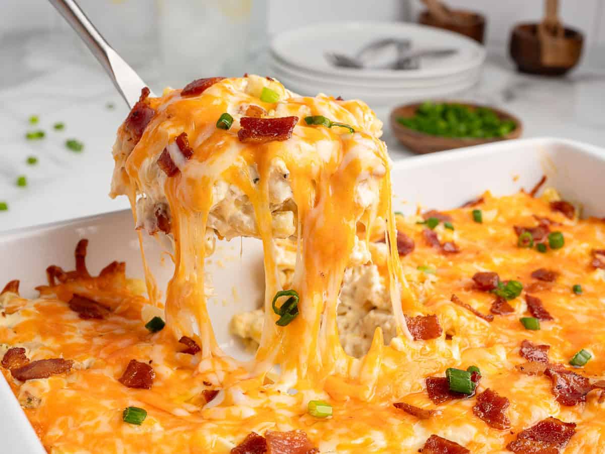 A cheesy Crack Chicken Casserole is being served from a white baking dish, with melted cheese stretching from the slice and topped with crispy bacon and chopped green onions. Plates and utensils are in the background, perfectly complementing this irresistible Crack Chicken Casserole Recipe.