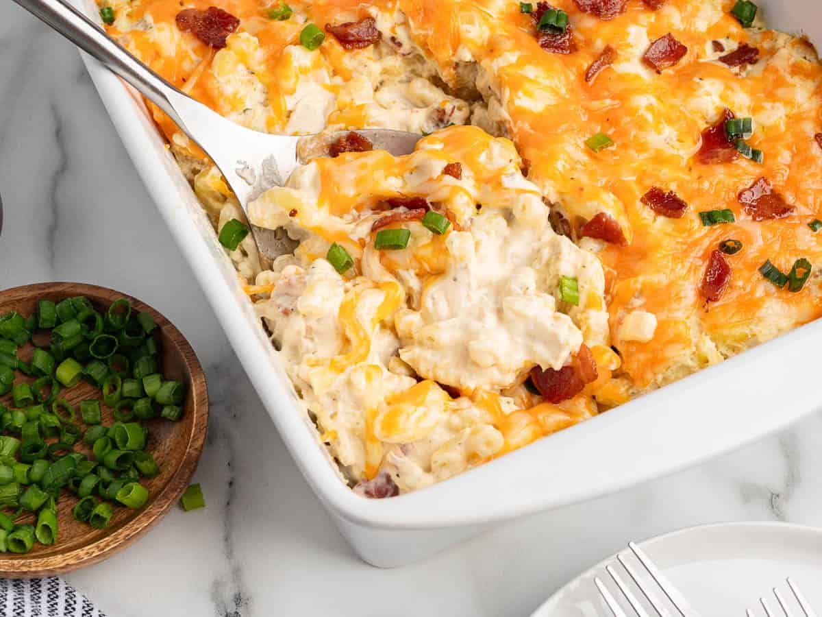 A cheesy Crack Chicken Casserole topped with bacon and green onions, with a serving being lifted out on a spatula. A small bowl of chopped green onions sits nearby on a marble countertop.