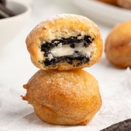 Two golden brown Deep Fried Oreos are stacked, with the top one cut open to reveal a creamy filling studded with dark cookie pieces. A light dusting of powdered sugar covers the crisp surface.