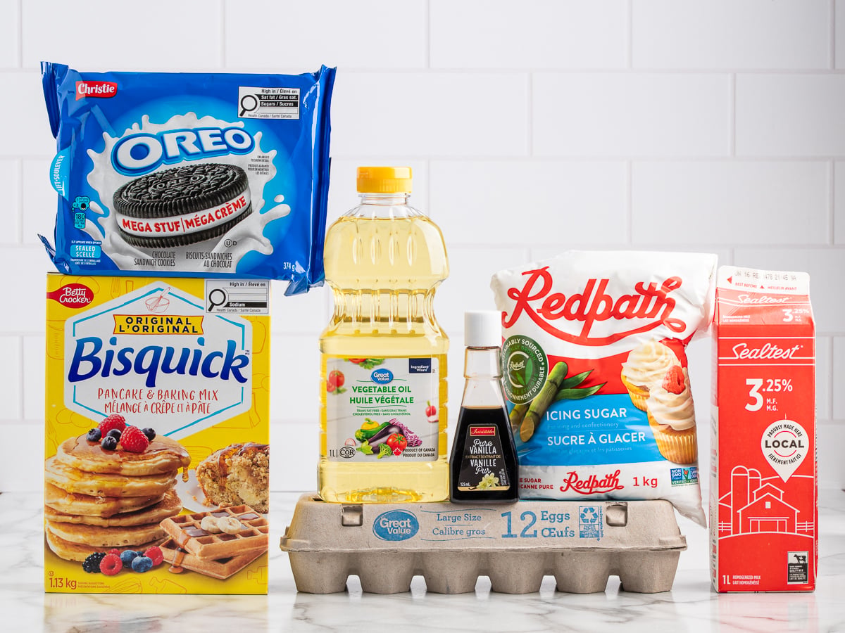 Assorted baking ingredients on a counter: Oreo cookies, Bisquick pancake mix, vegetable oil, vanilla extract, icing sugar, milk in a carton, and eggs—everything you need to make delicious Deep Fried Oreos—against a white tiled background.