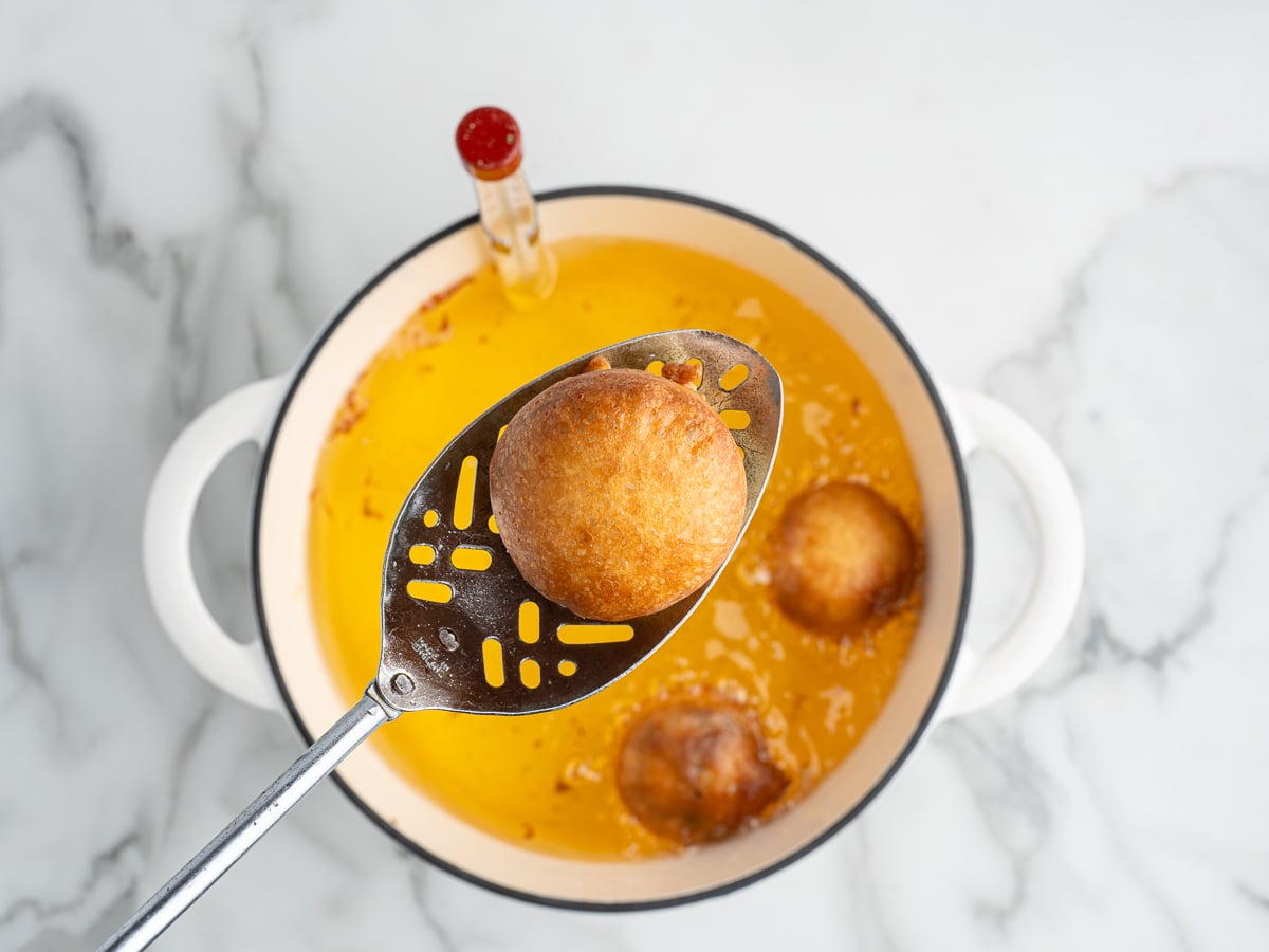 A slotted spoon holds a golden-brown Deep Fried Oreo above a white pot of hot oil, with more Oreos frying inside and a thermometer clipped to the pot on a marble surface.