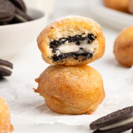 A stack of Deep Fried Oreos, with the top one cut open to reveal a creamy cookie center. Other whole cookies and a bowl of cookies sit in the background. The treats are generously dusted with powdered sugar.