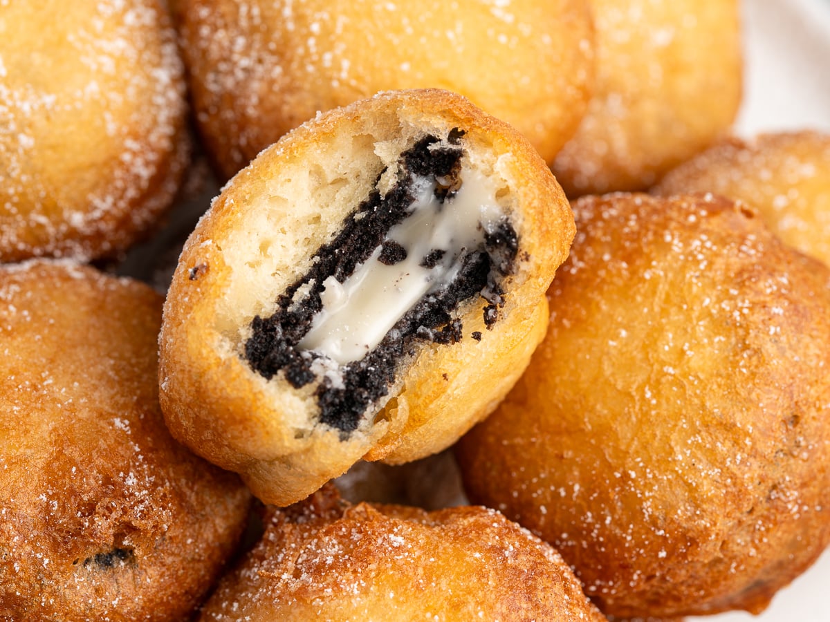 Close-up of several golden, sugar-dusted Deep Fried Oreos, with one broken open to reveal a creamy, black-and-white filling inside, showcasing the cookie’s soft interior and melted center.