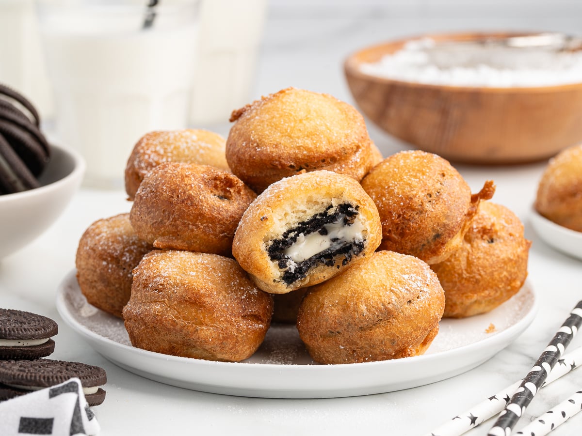A plate piled with golden brown Deep Fried Oreos, one cut open to reveal the chocolate cookie and cream filling inside. Glasses of milk and extra Oreos are visible in the background.