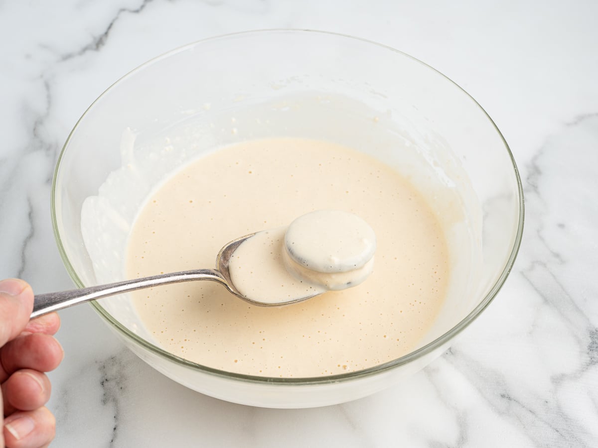 A hand holds a spoon dipping a round slice of banana into a bowl of light, creamy batter—perfect for making treats like Deep Fried Oreos—on a marble countertop.