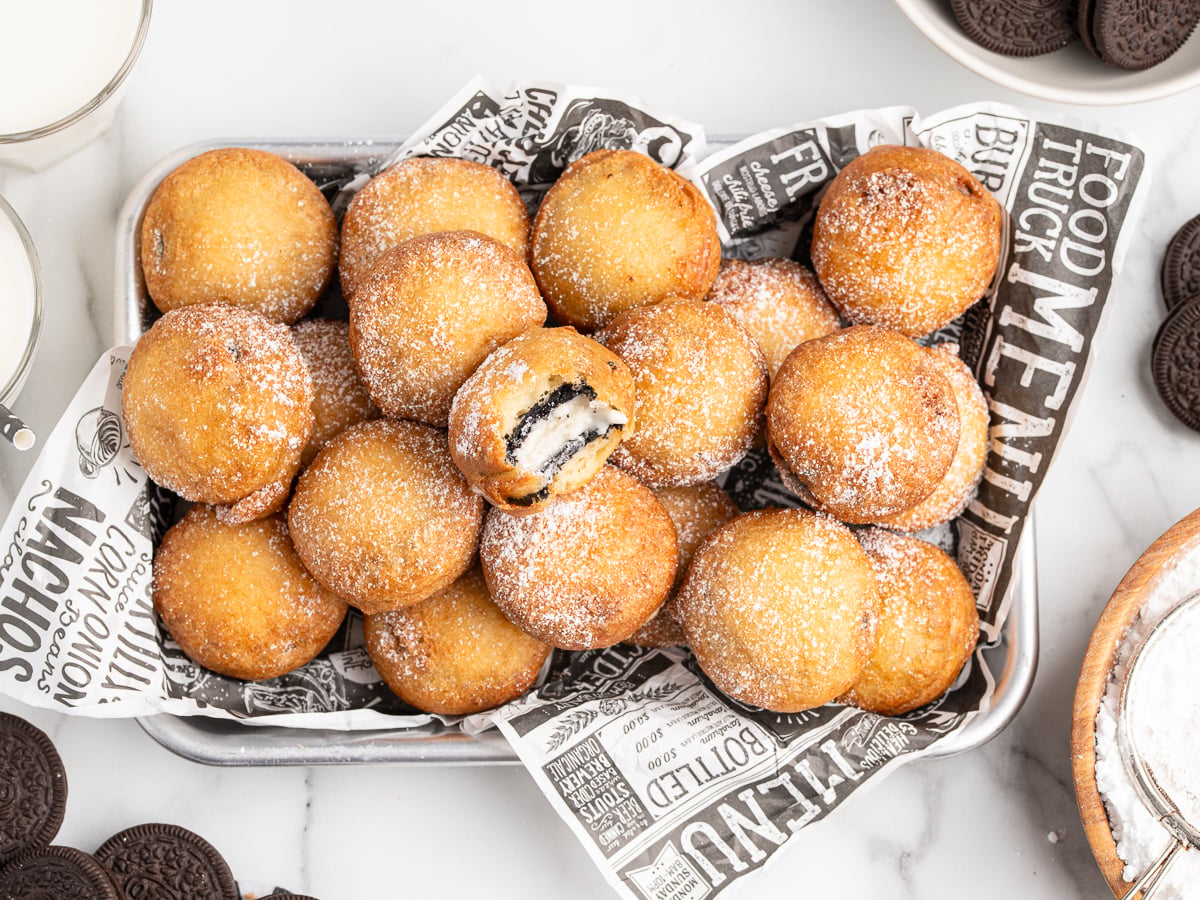 A tray lined with decorative paper holds Deep Fried Oreos—golden brown treats dusted with powdered sugar. One is cut open to reveal a chocolate cookie inside, with extra Oreo cookies and glasses of milk nearby.