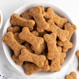 Bowl filled with homemade bone-shaped peanut butter dog treats on a white surface.
