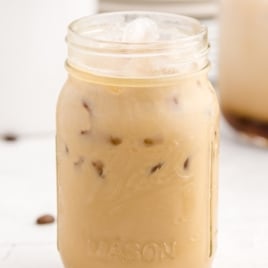 A mason jar filled with Iced Caramel Macchiato sits on a white surface. Ice cubes glisten in the light brown drink, coffee beans are scattered nearby, and another glass of this Iced Caramel Macchiato recipe is blurred in the background.