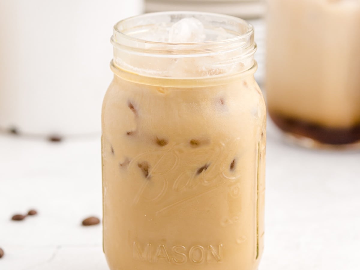 A mason jar filled with Iced Caramel Macchiato sits on a white surface. Ice cubes glisten in the light brown drink, coffee beans are scattered nearby, and another glass of this Iced Caramel Macchiato recipe is blurred in the background.