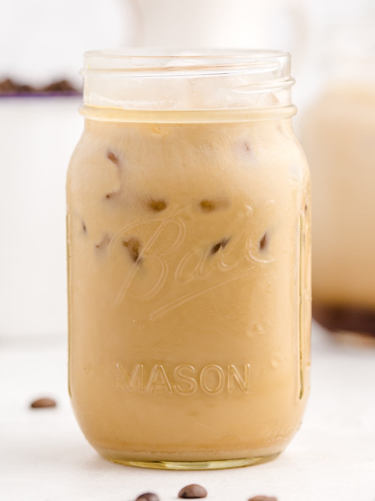 A mason jar labeled Ball Mason is filled with a refreshing Iced Caramel Macchiato and ice cubes, sitting on a light surface with coffee beans scattered nearby—perfect inspiration for your next Iced Caramel Macchiato recipe.