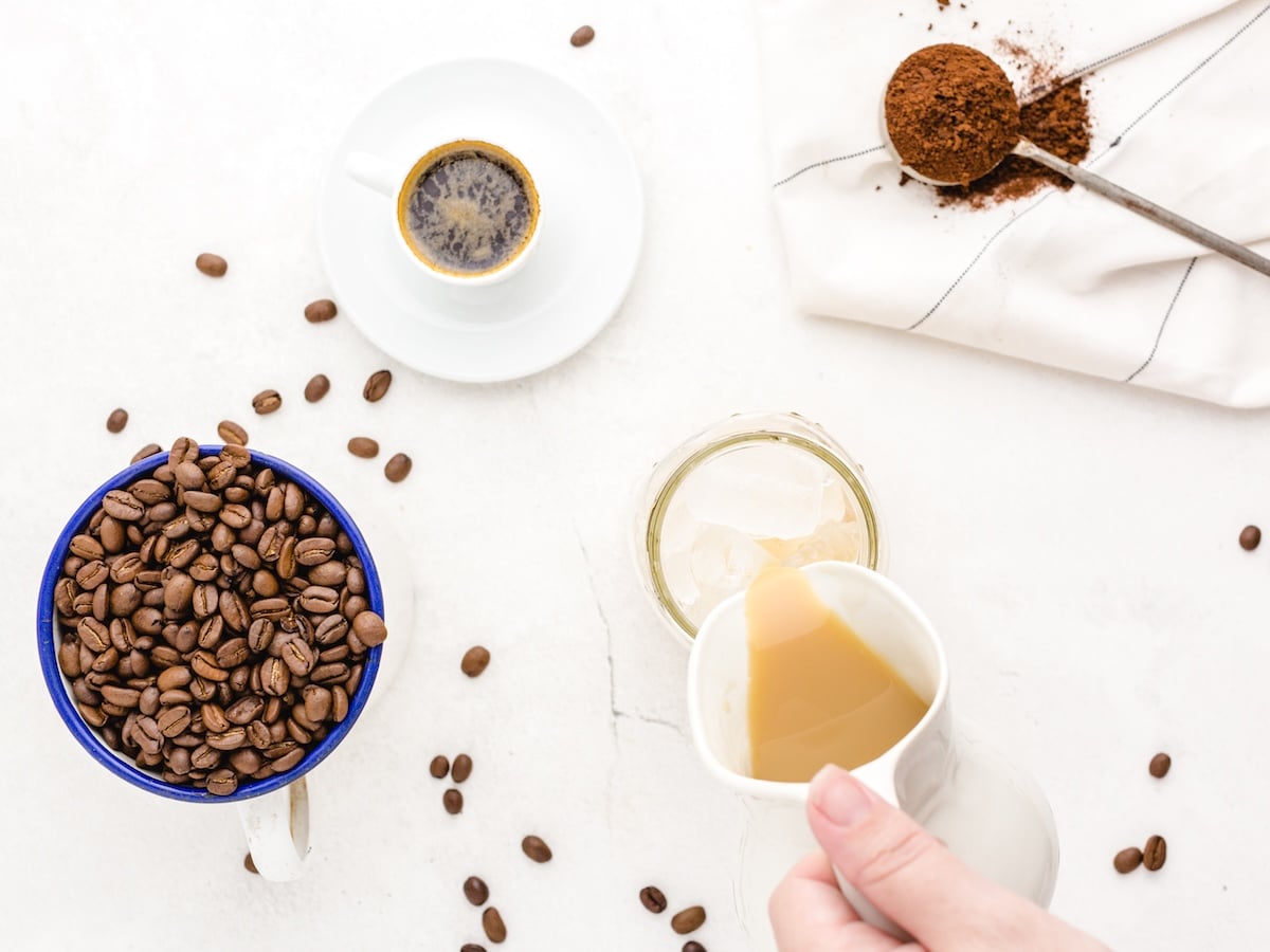A hand pours milk into a glass with ice, surrounded by a cup of coffee beans, black coffee, a spoon with ground coffee, and scattered beans—perfect inspiration for your next Iced Caramel Macchiato or homemade Iced Caramel Macchiato recipe.