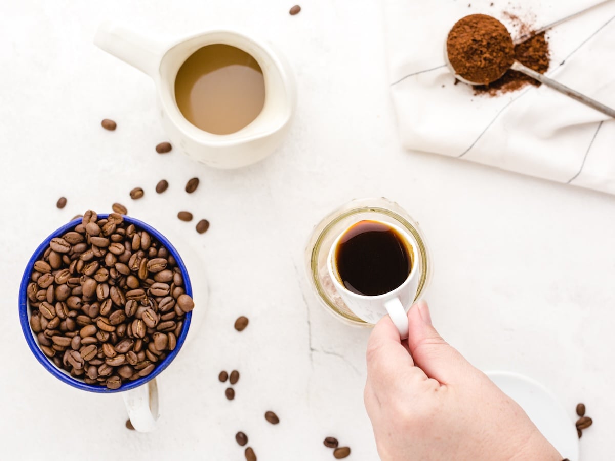 A hand holds a cup of black coffee on a white surface. Nearby are a mug of beans, creamer, scattered beans, and a spoon with grounds—perfect inspiration for your next Iced Caramel Macchiato Recipe.