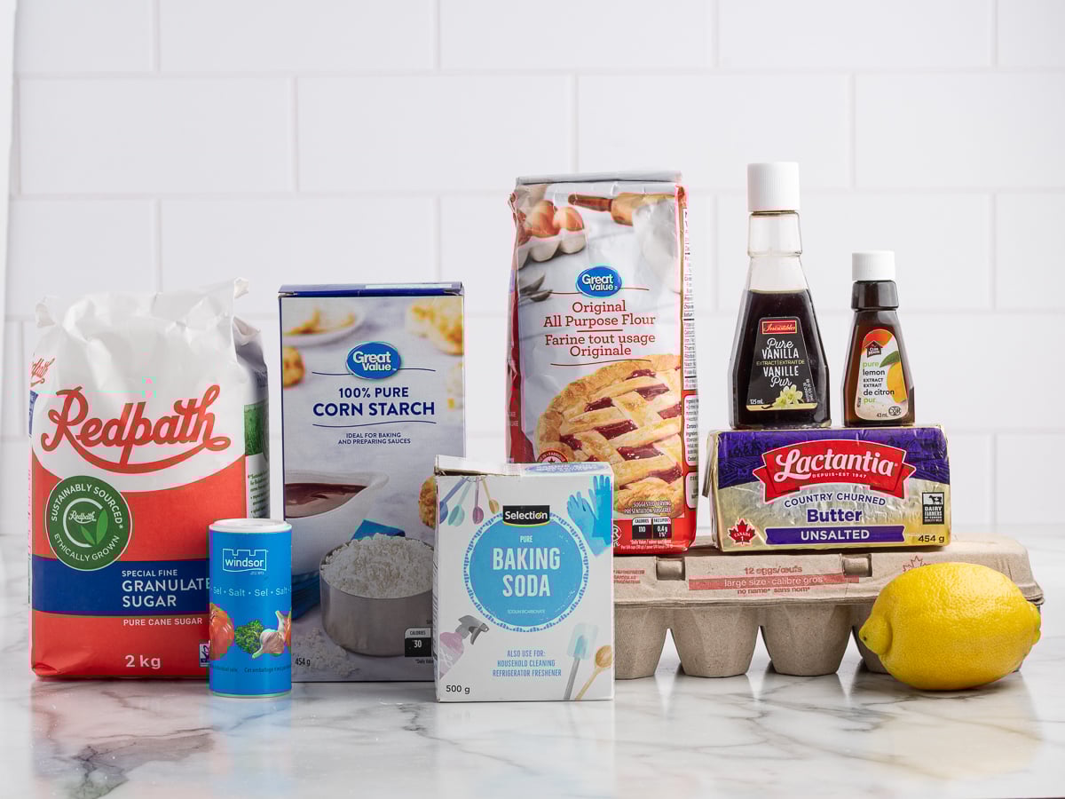 A collection of baking ingredients for Lemon Sugar Cookies on a counter&mdash;sugar, salt, cornstarch, flour, baking soda, butter, eggs, vanilla extract, artificial vanilla flavor, and a lemon&mdash;arranged in front of a white tile wall.