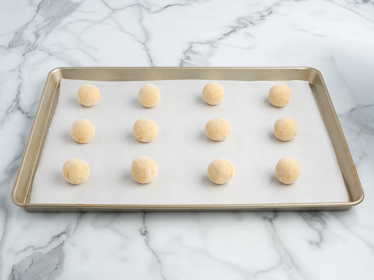 A baking sheet lined with parchment paper holds twelve evenly spaced, round Lemon Sugar Cookies dough balls, placed in a 3 by 4 grid on a marble countertop.