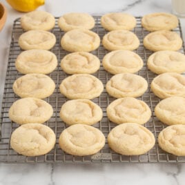 A batch of round, golden Lemon Sugar Cookies cools on a wire rack. Slices of lemon, a bowl of lemons, and glasses of milk are nearby on a white marble surface.