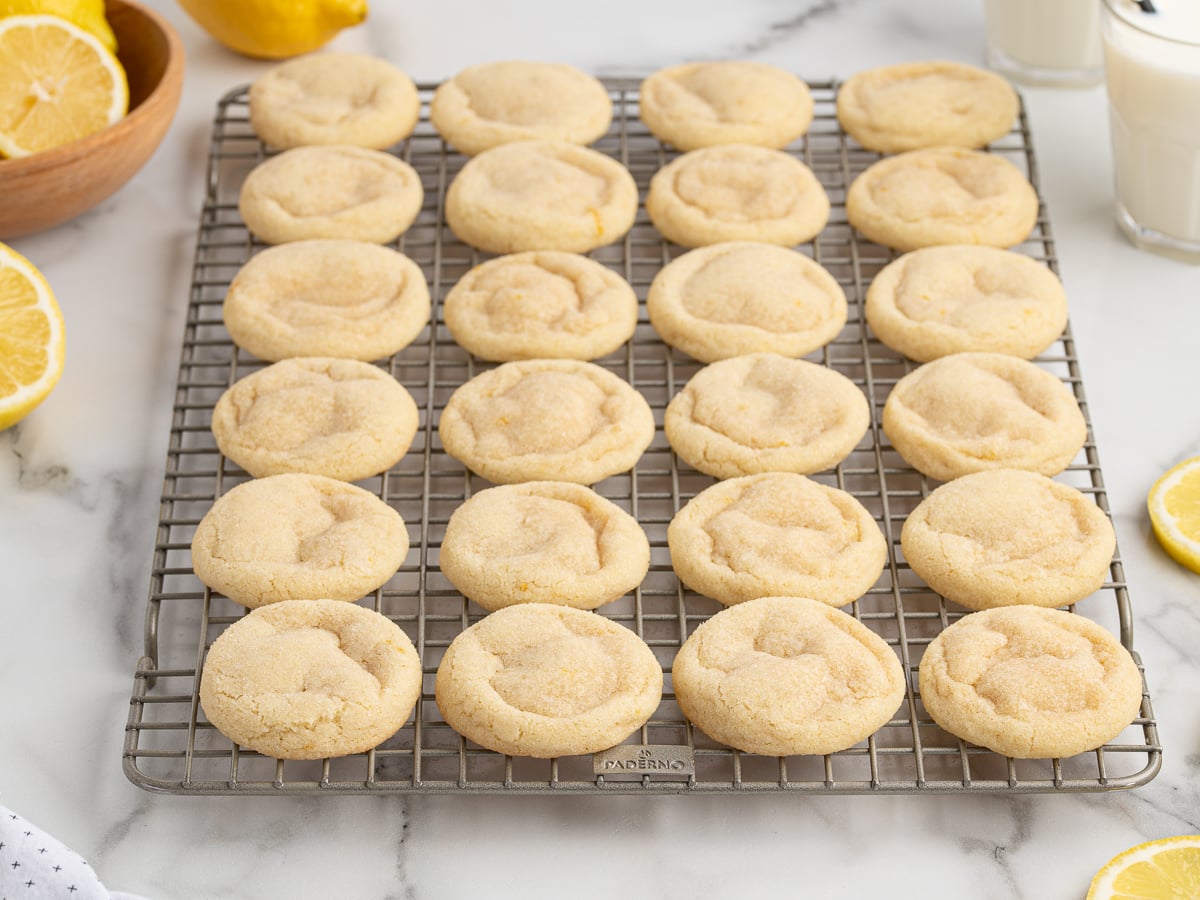 A batch of round, golden Lemon Sugar Cookies cools on a wire rack. Slices of lemon, a bowl of lemons, and glasses of milk are nearby on a white marble surface.