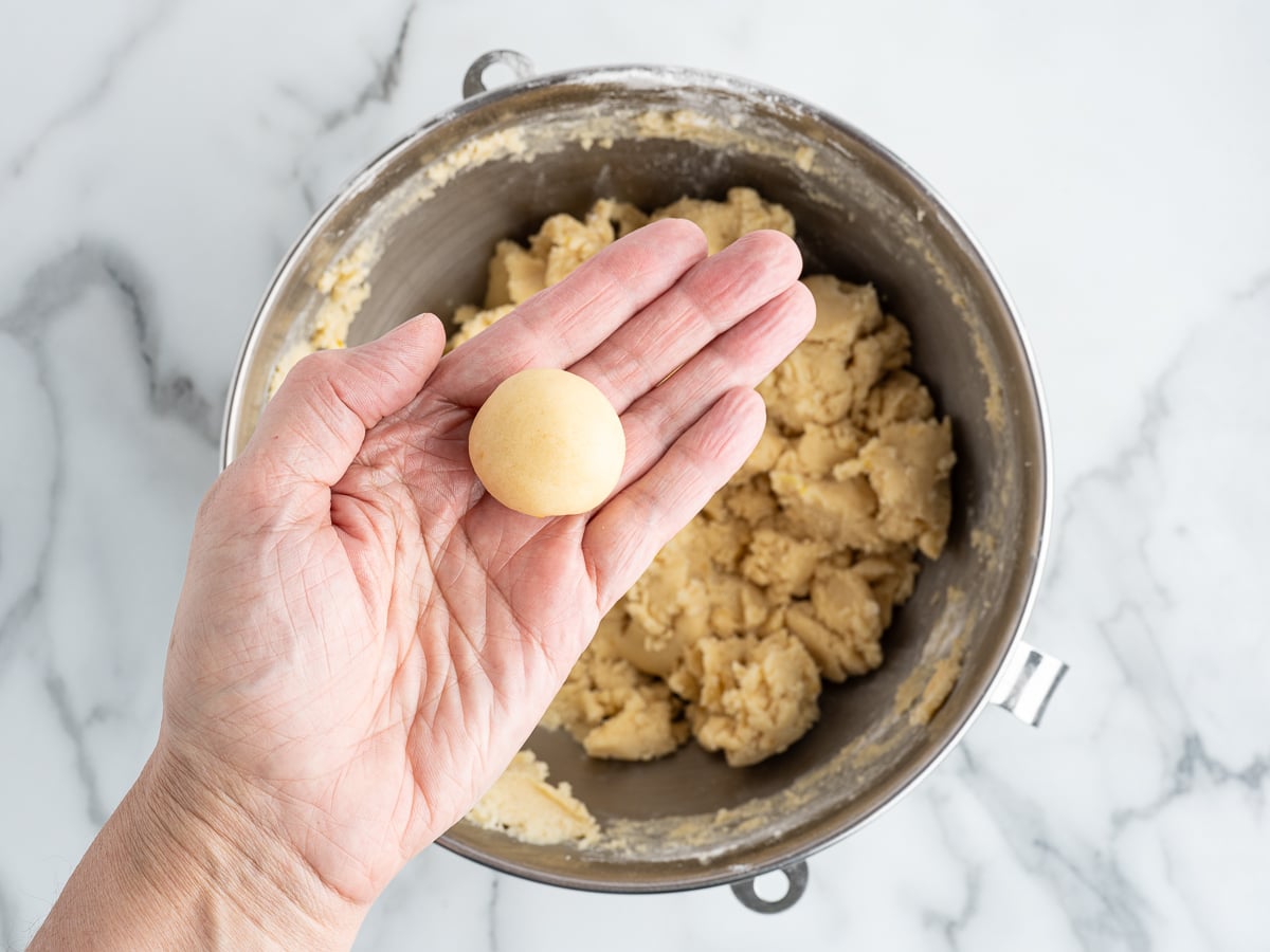 A hand holds a small, round ball of cookie dough above a metal mixing bowl filled with more dough on a marble surface, ready to bake delicious Lemon Sugar Cookies.