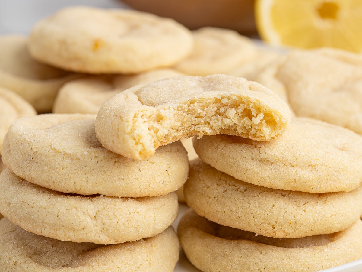 A close-up of stacked lemon sugar cookies, made with our favorite lemon cookie recipe, with one cookie on top showing a bite taken out. The cookies look soft and chewy, and a fresh lemon slice is visible in the background.