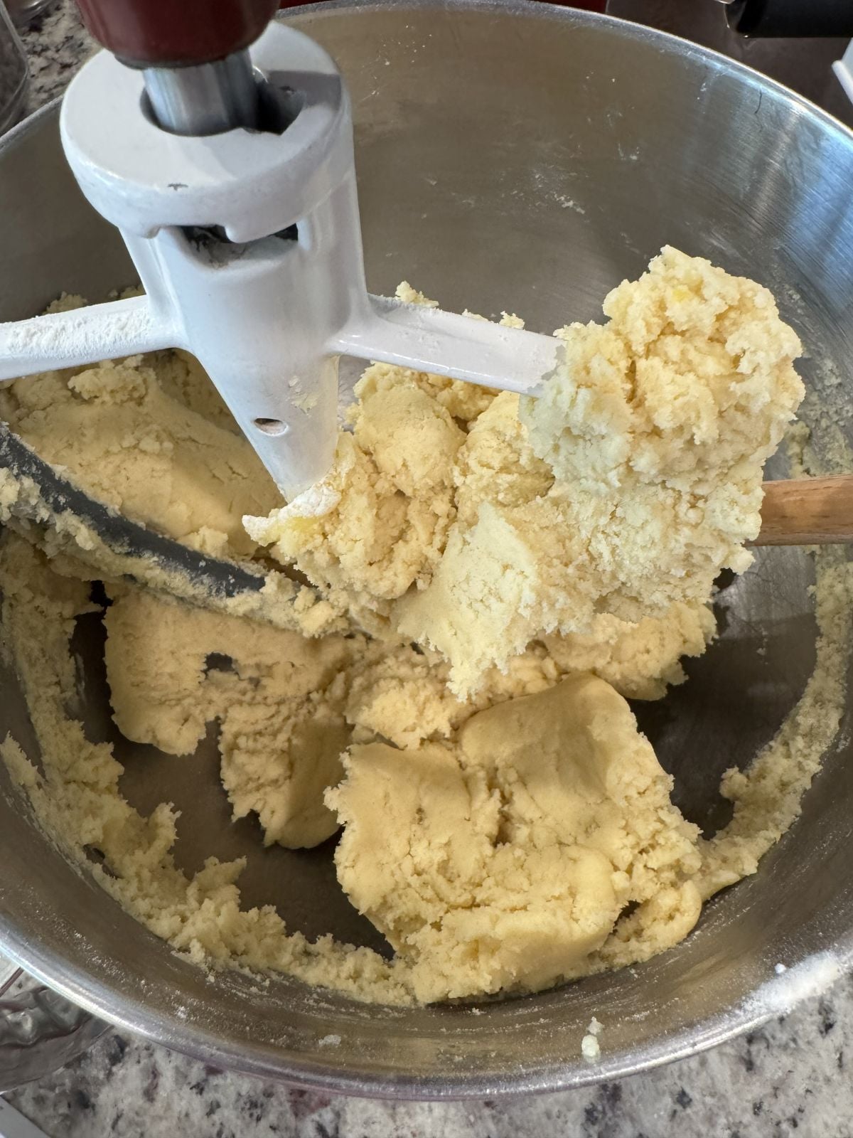 A stand mixer with a paddle attachment is blending thick, pale yellow dough for a Lemon Cookie Recipe in a metal bowl. Some dough clings to the paddle, and a wooden spoon rests inside, ready to help shape delicious Lemon Sugar Cookies.