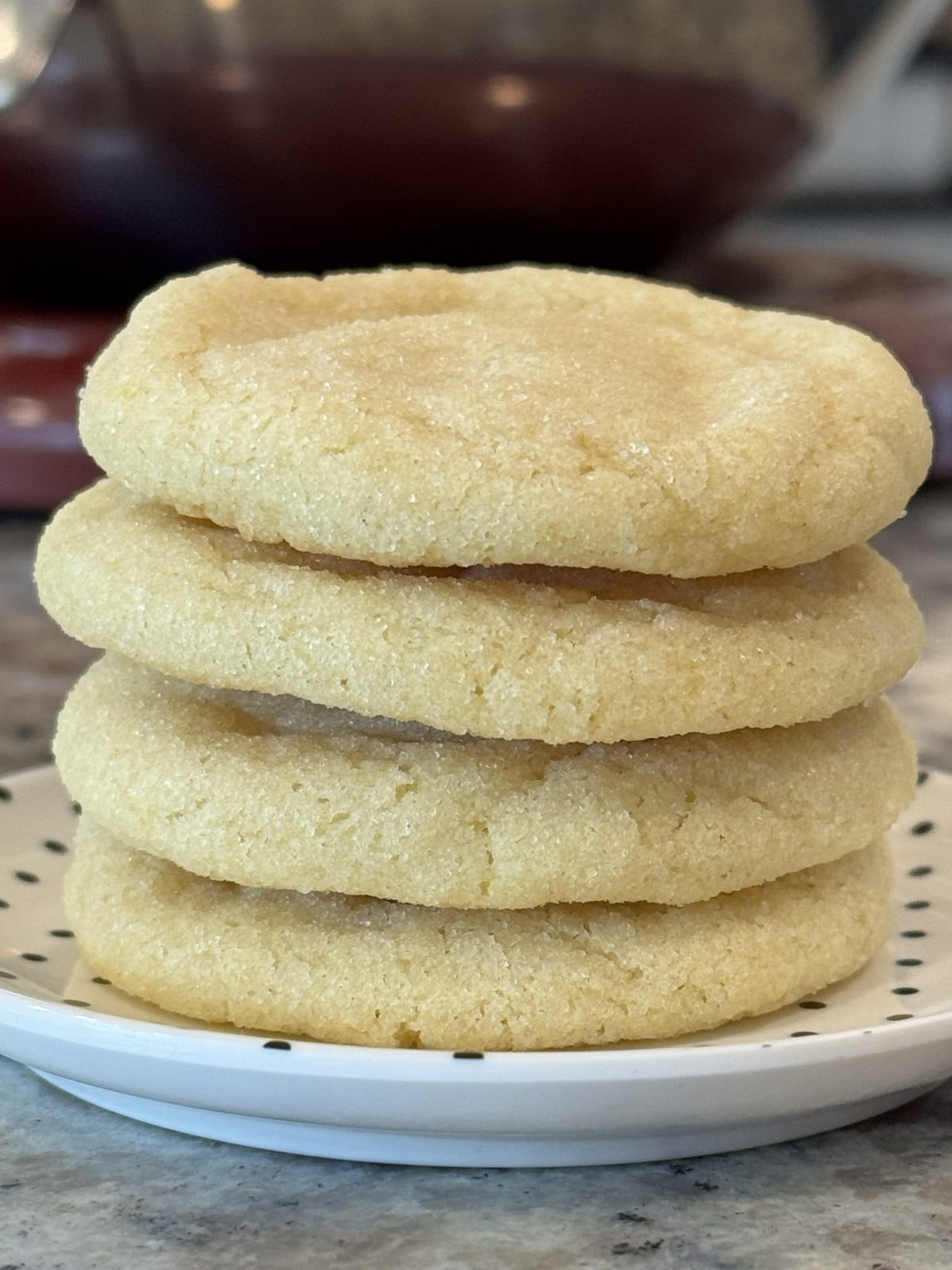 A stack of four lemon sugar cookies sits on a white plate with black dots, with a blurred background showing a marble countertop and a mixing bowl&mdash;perfect inspiration for your next lemon cookie recipe.