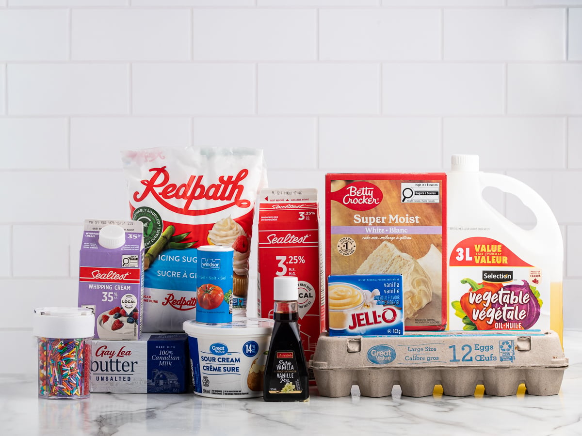 A variety of baking ingredients for vanilla cupcakes on a counter, including eggs, sour cream, milk, butter, vanilla extract, whipped cream, cake mix, sugar, sprinkles, pudding mix, and vegetable oil against a white tile background.