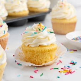 A Vanilla Cupcakes sits on a marble surface, topped with swirled white frosting and colorful sprinkles, its wrapper partly unwrapped. More vanilla cupcakes and scattered sprinkles surround this delightful treat.