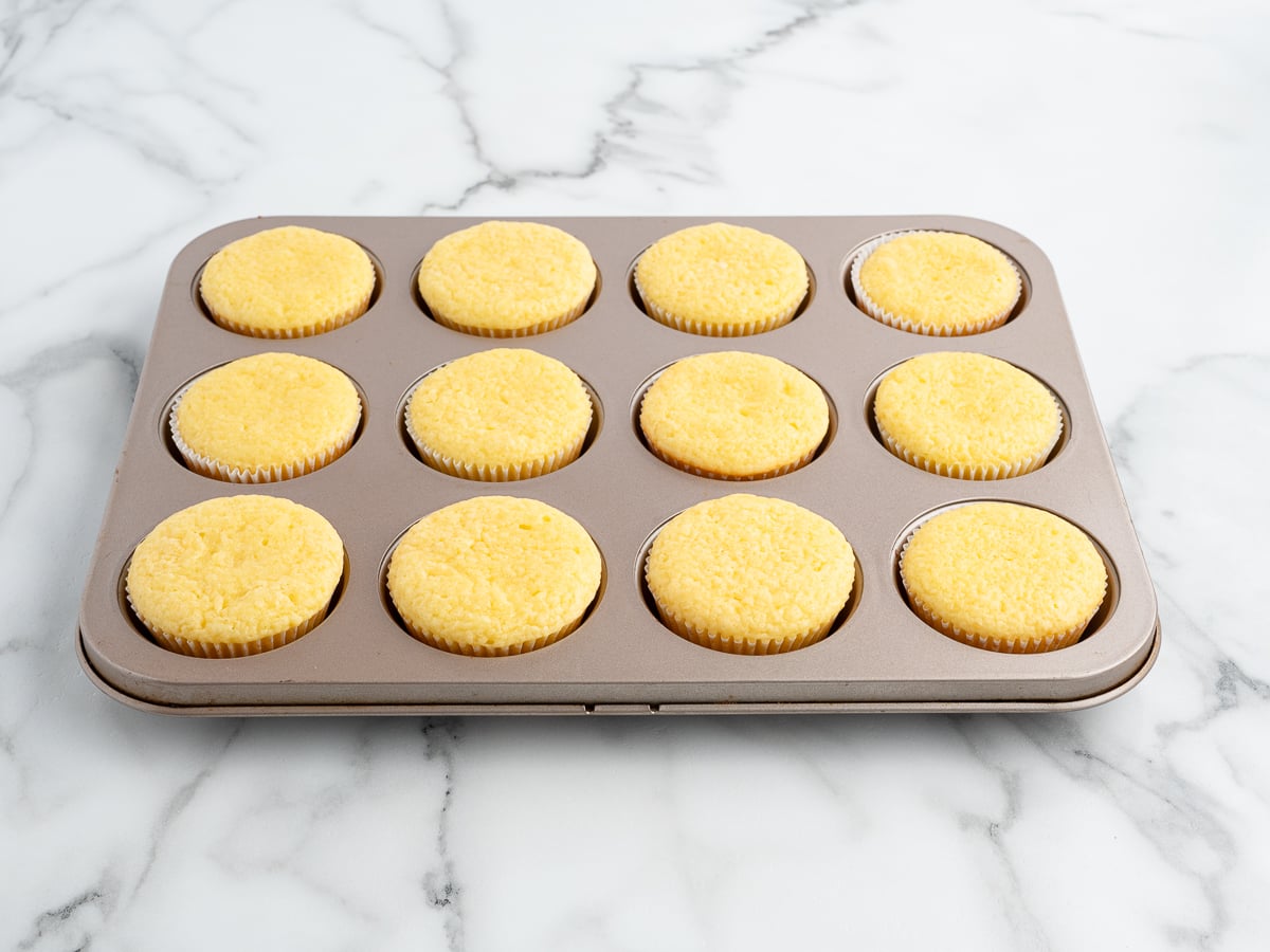 A muffin tin with twelve freshly baked vanilla cupcakes sits on a white marble countertop.