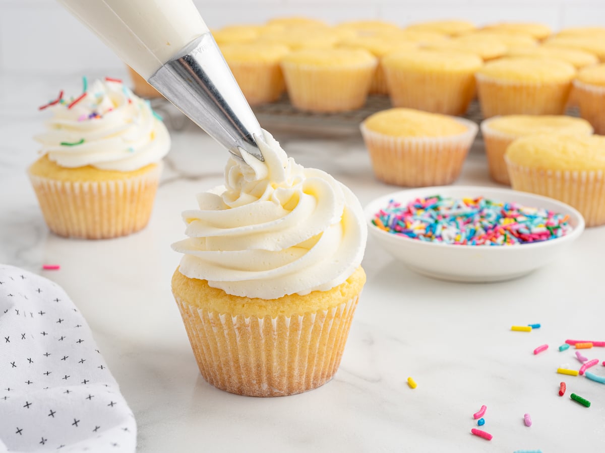 A batch of Vanilla Cupcakes is being decorated as one cupcake gets topped with white icing from a piping bag. Nearby, unfrosted cupcakes, a bowl of colorful sprinkles, and a white napkin rest on the marble surface.
