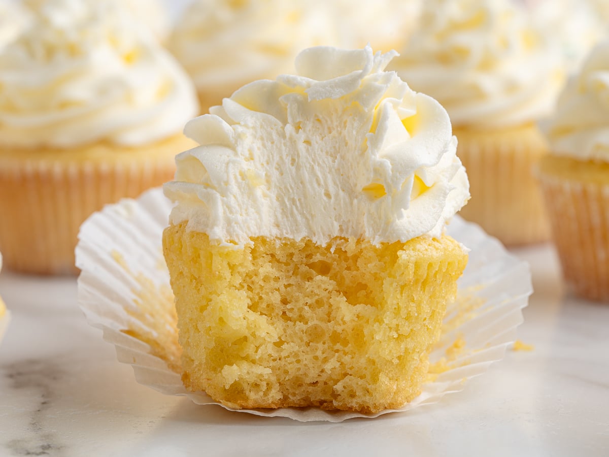 A yellow Vanilla Cupcake with fluffy white frosting sits on an opened paper liner. The cupcake has a large bite taken out, revealing its soft, airy interior. More frosted Vanilla Cupcakes are blurred in the background.