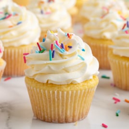 A close-up of Vanilla Cupcakes topped with swirled white frosting and colorful rainbow sprinkles, beautifully arranged on a white surface.