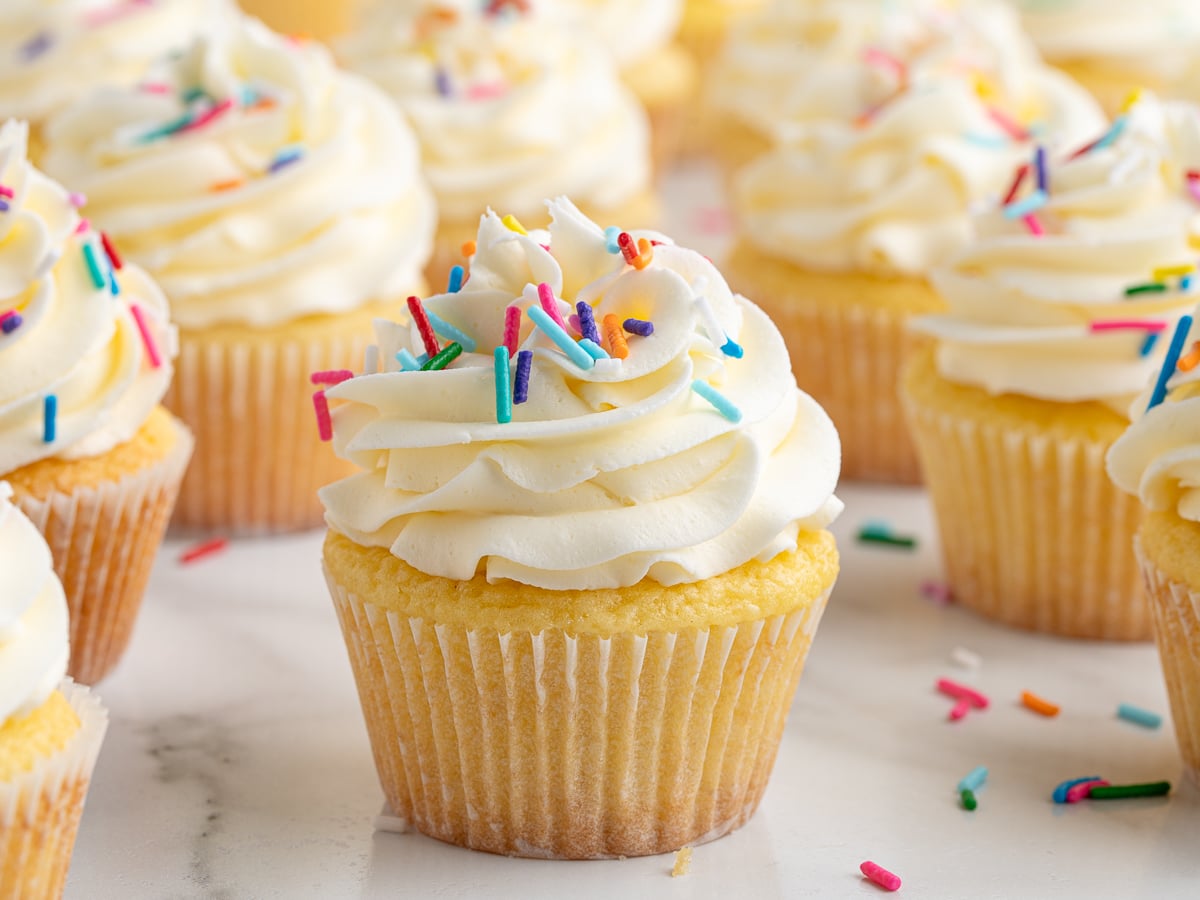 A close-up of Vanilla Cupcakes topped with swirled white frosting and colorful rainbow sprinkles, beautifully arranged on a white surface.