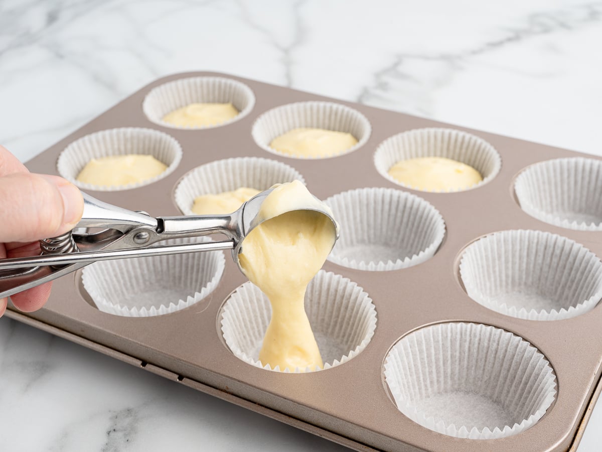 A hand uses an ice cream scoop to fill a paper-lined muffin tin with vanilla cupcakes batter on a marble countertop.