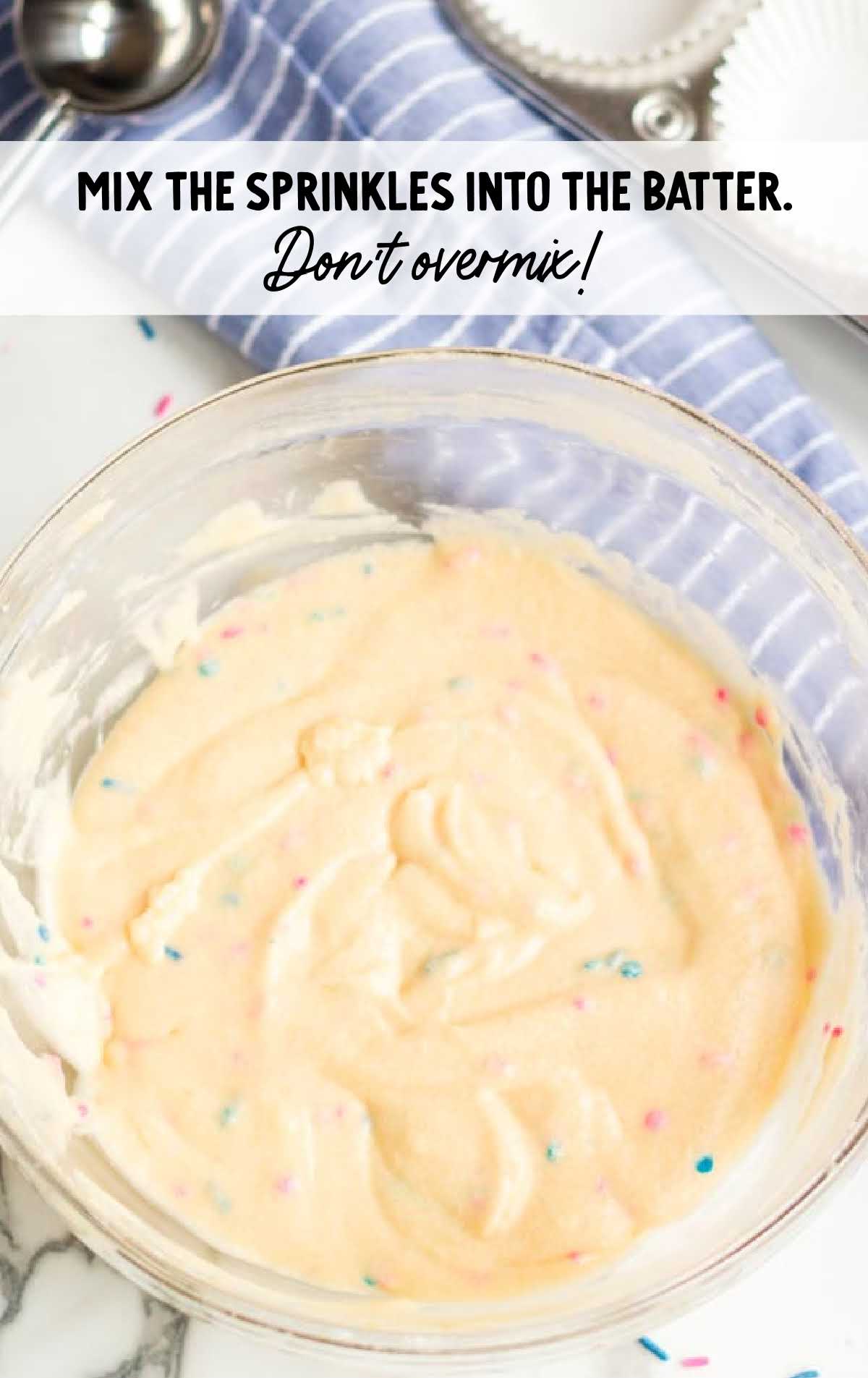 Mixing bowl of funfetti cupcake batter with pink and blue sprinkles being folded in, alongside cupcake liners and baking tools.
